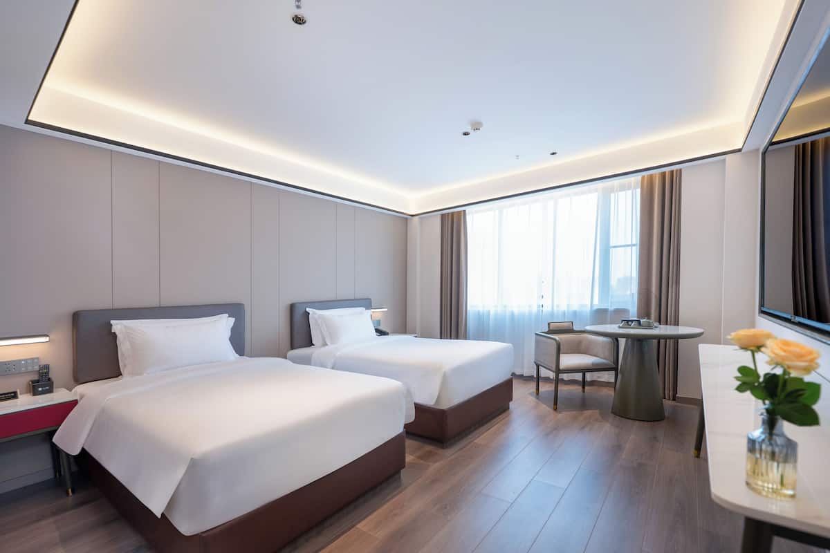 A hotel room with two beds, a desk, a chair, a round table, and a vase with flowers.