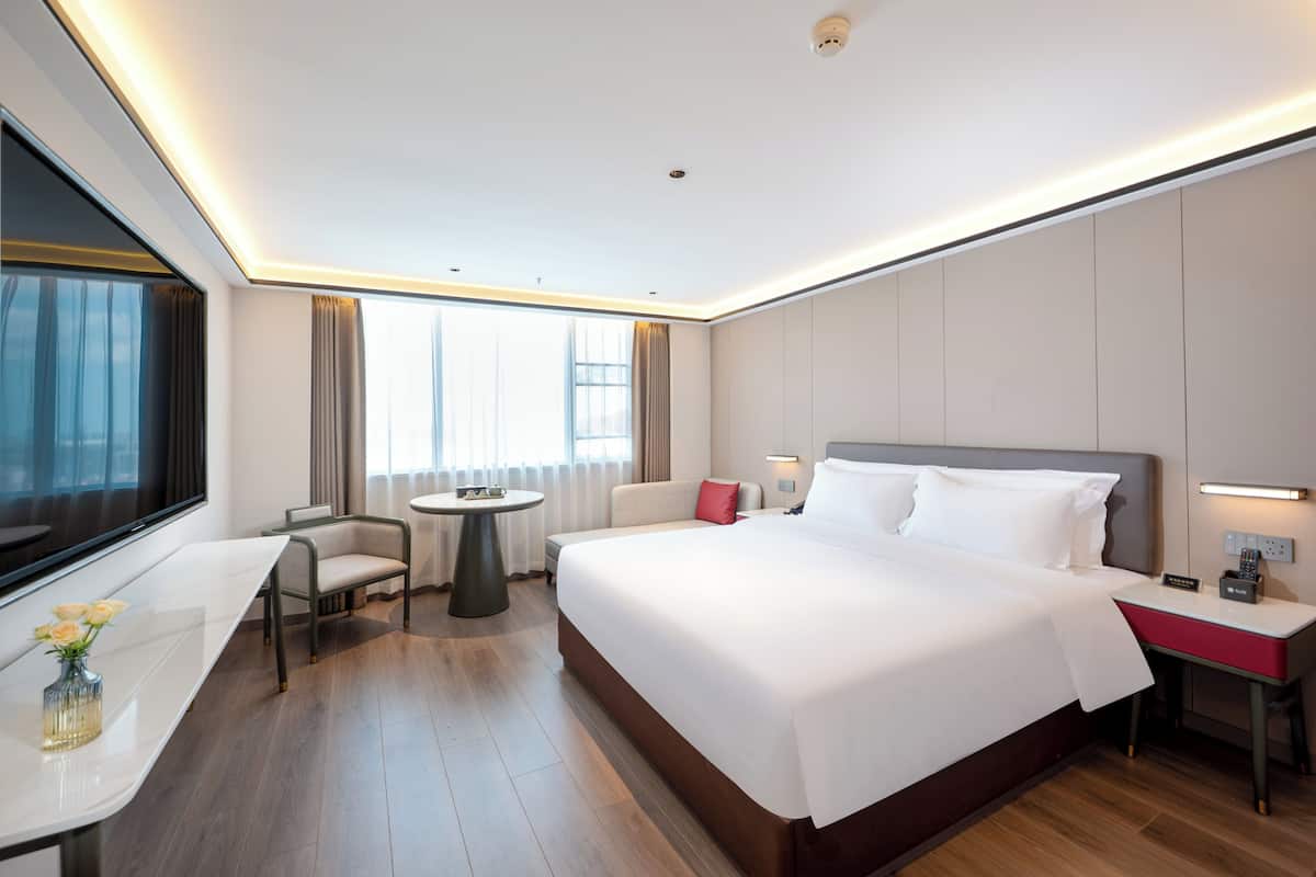 A modern hotel room with a large bed, a desk, a chair, a small table, and a window with curtains.