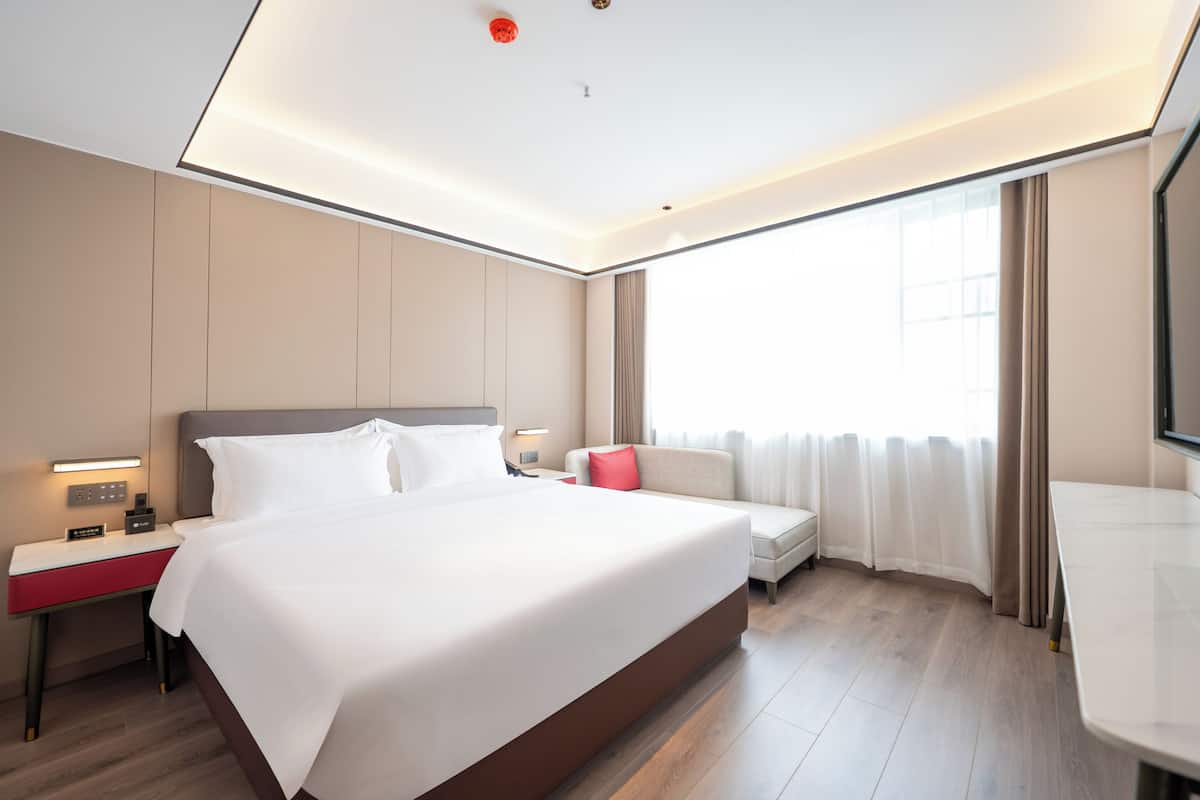 A modern hotel room with a large bed, bedside tables, a desk, a sofa, and a large window with curtains.