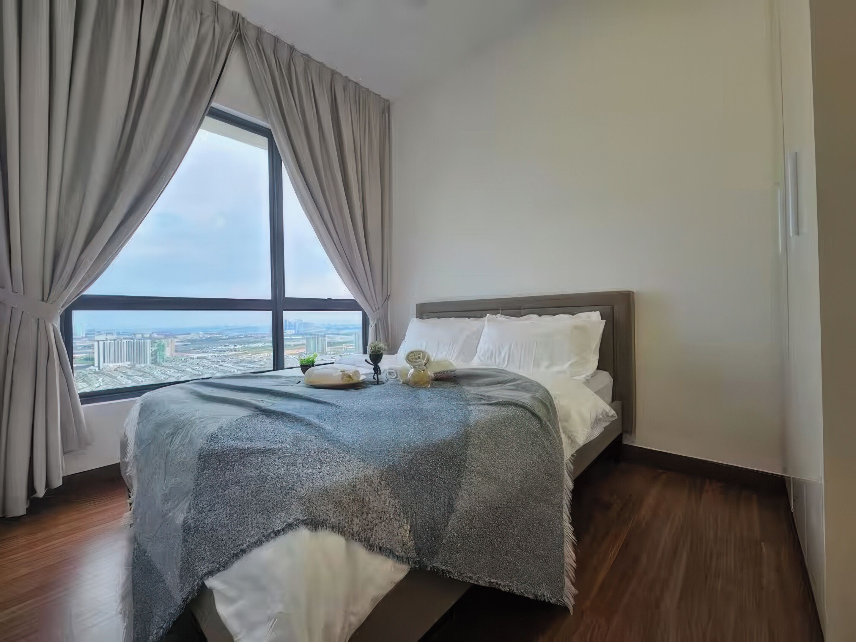 A bedroom with a large window, a bed with a grey blanket, and a view of a cityscape.