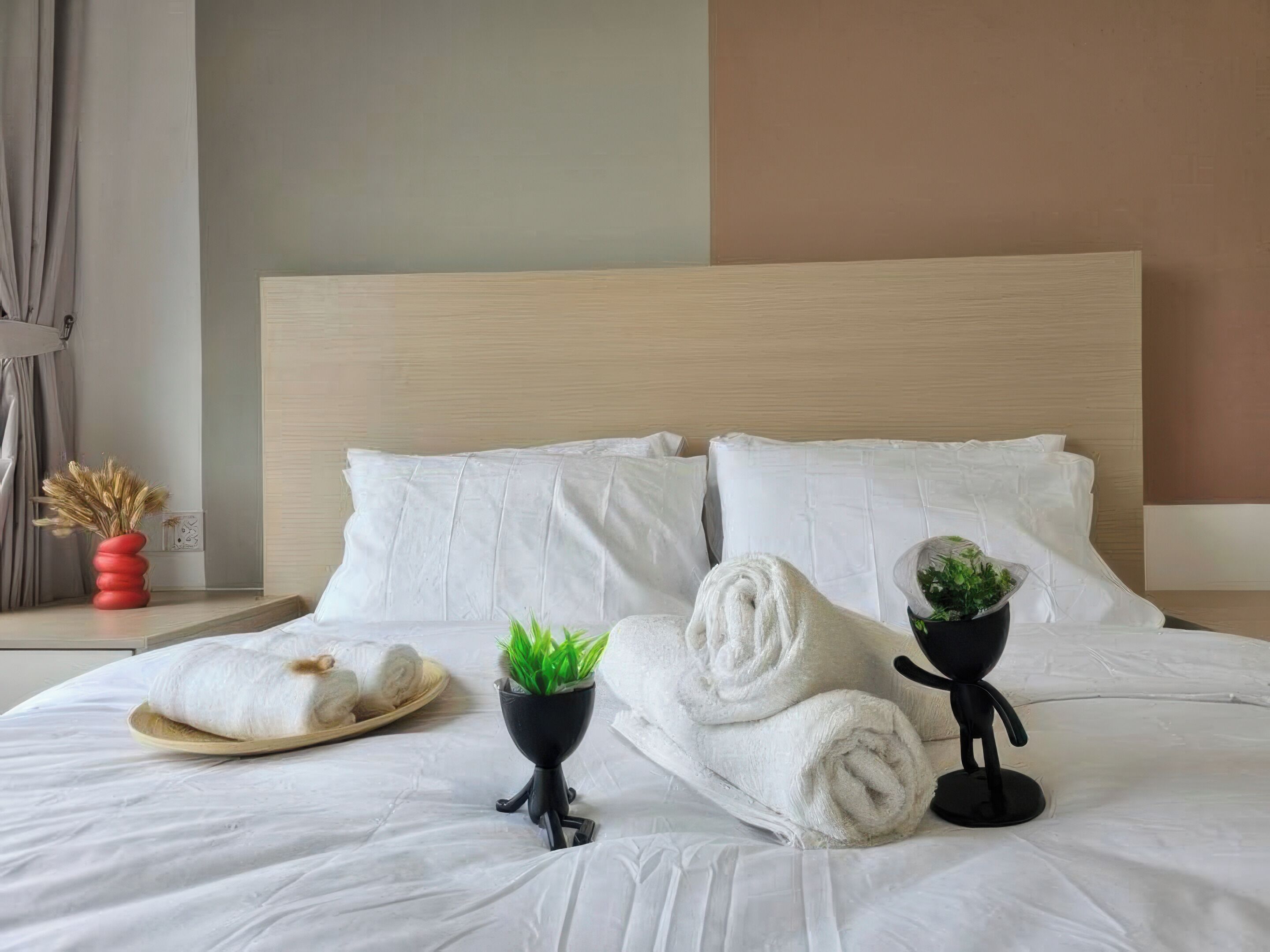 Una cama bien tendida con sábanas blancas, un florero rojo con plantas secas, una maceta negra con plantas verdes y toallas enrolladas.