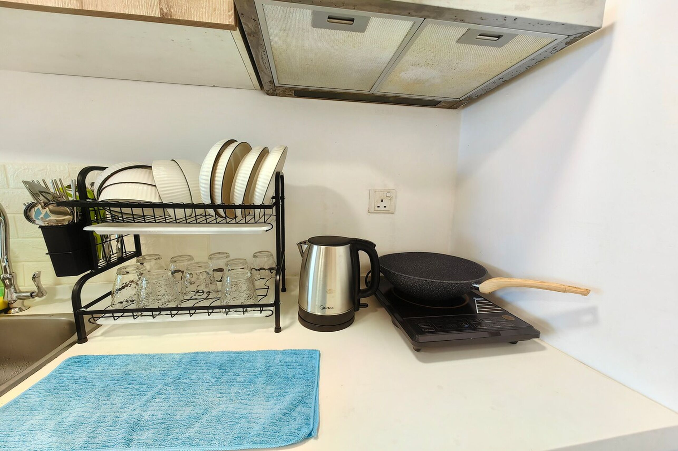 A kitchen with a dish rack holding plates and glasses, a kettle, a frying pan, and a blue towel on the counter.