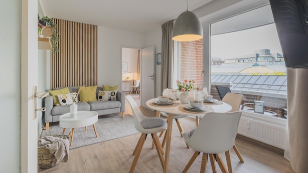 A modern living room with a sofa, dining table, chairs, and a large window with a view of a building. - Rentaflat Norderstedt 2BR Apartment for 4 Guests (Norderstedt)
