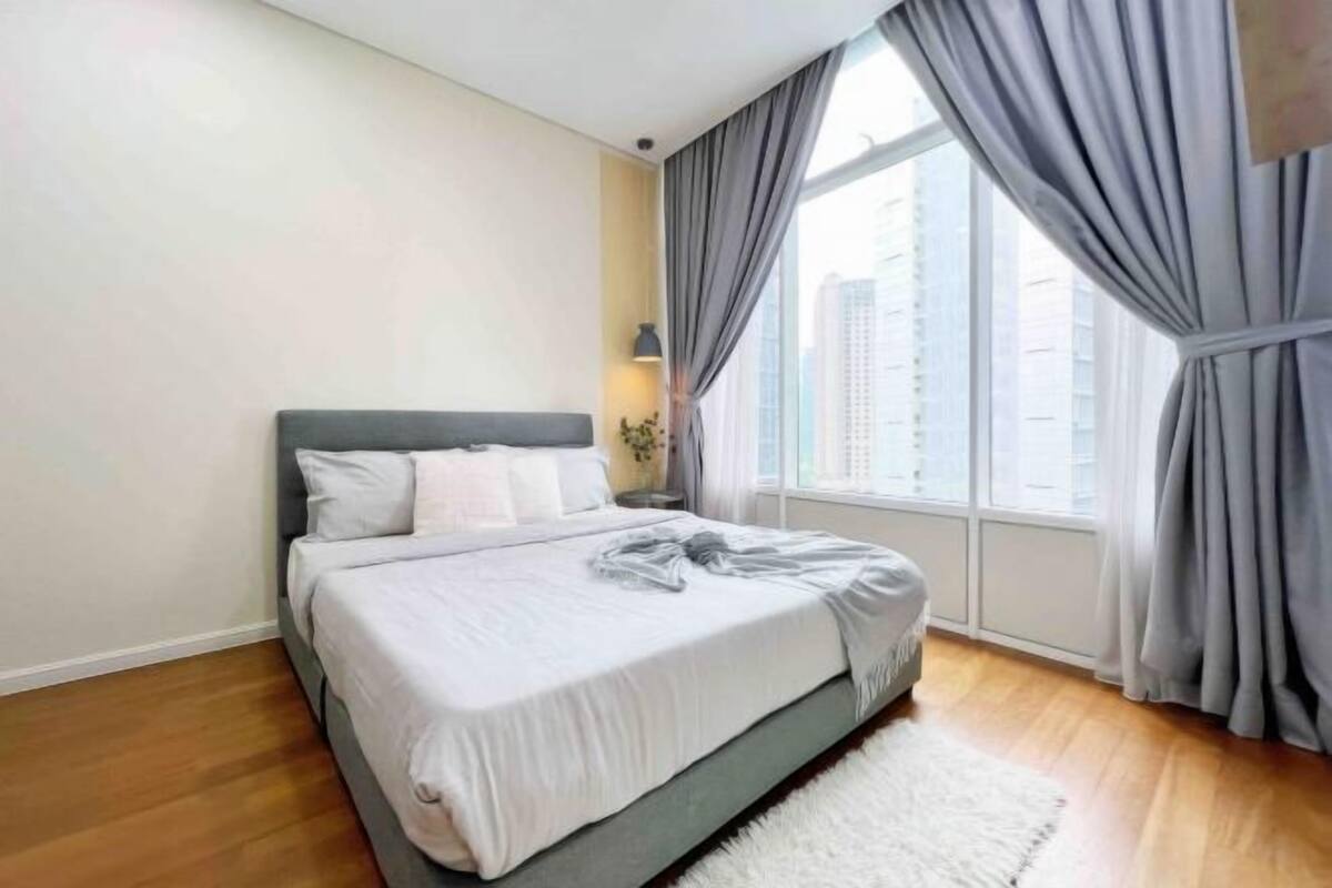 A bedroom with a bed, a window with curtains, a lamp, and a view of buildings.