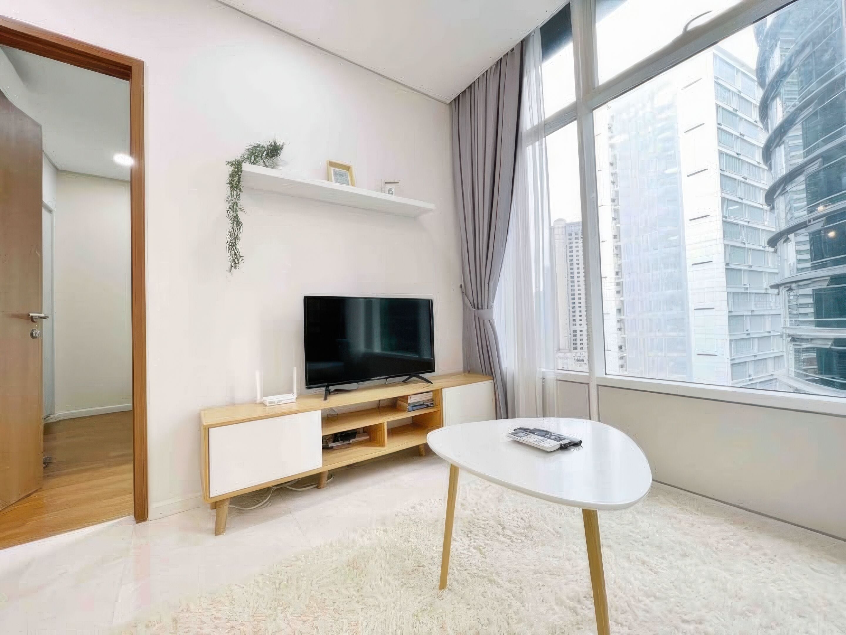 Una sala de estar moderna con un televisor de pantalla plana sobre un soporte de madera, una pequeña mesa blanca y un gran ventanal con vista a la ciudad.
