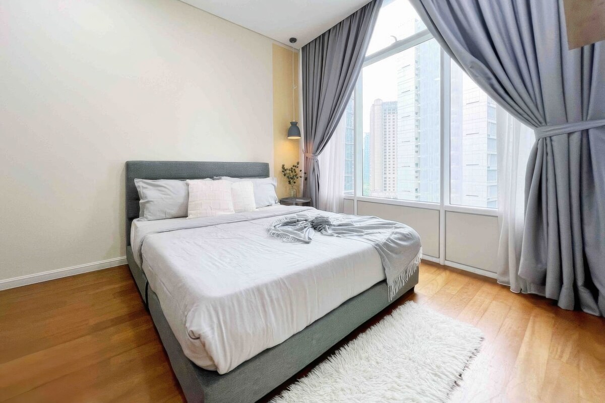 A modern bedroom with a bed, a nightstand, a lamp, and a view of buildings through the window.
