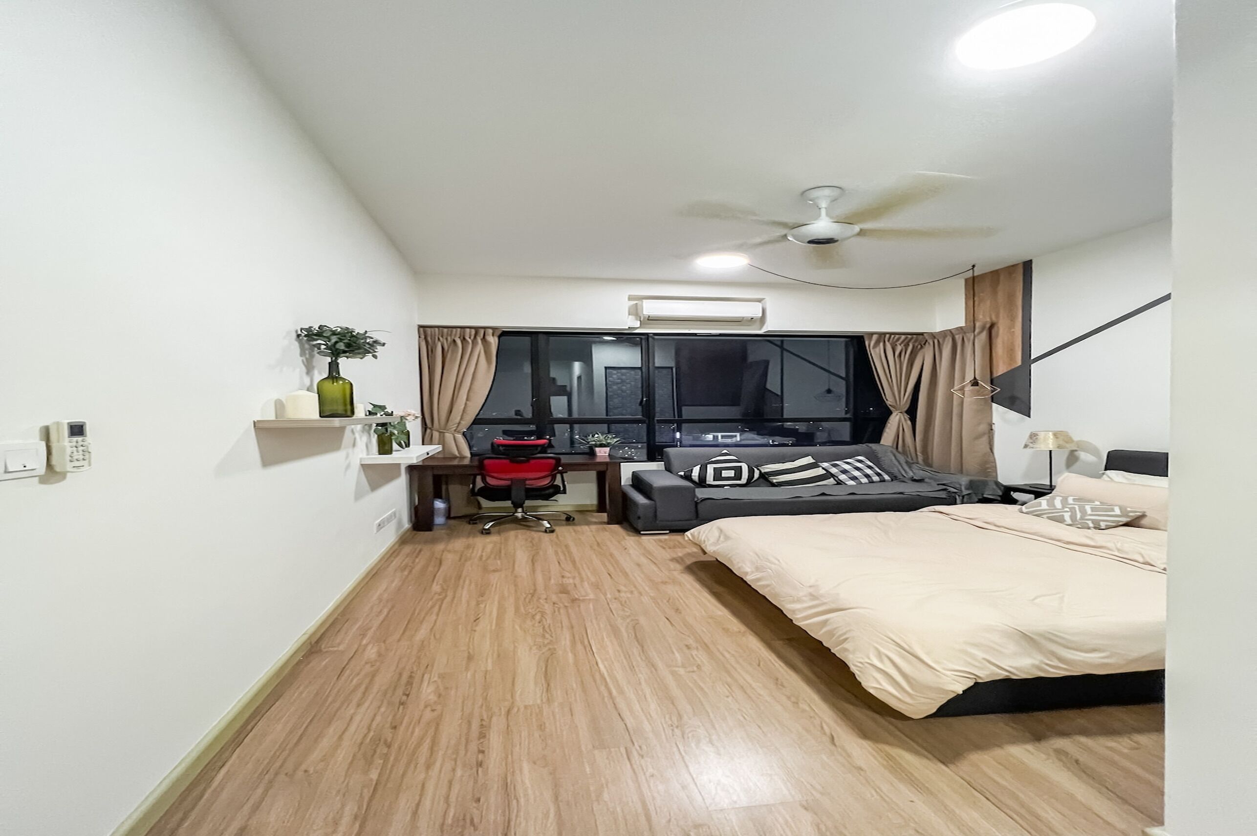 Ein kompaktes Hotelzimmer mit Bett, Schreibtisch mit Stuhl, kleinem Tisch, Fenster mit Vorhängen, Deckenventilator und Klimaanlage.
