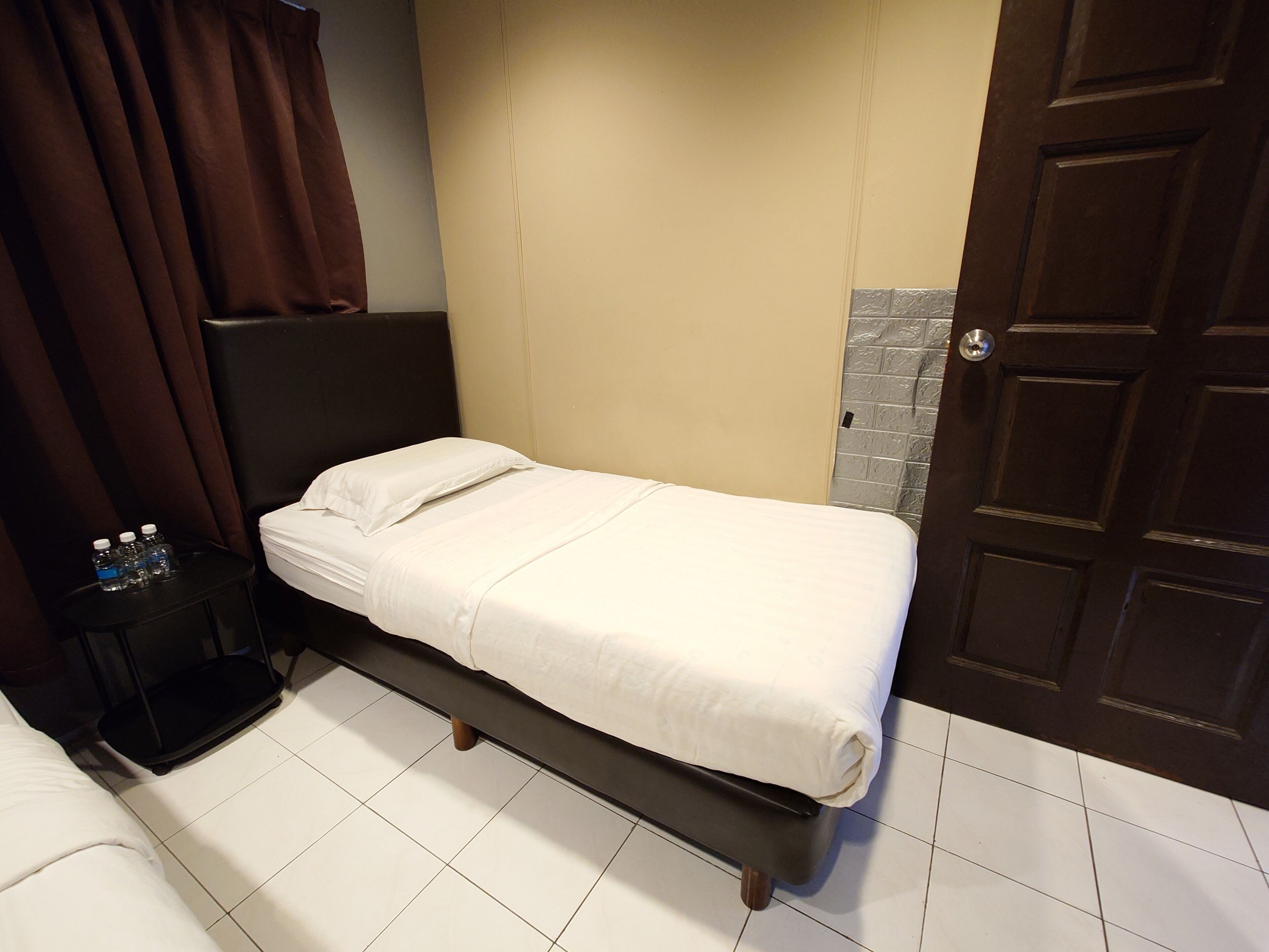 A single bed with a white mattress and pillows, a black headboard, a small bedside table with water bottles, and a door with a textured panel.