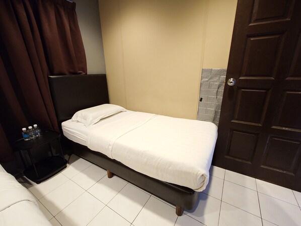 A single bed with a white mattress and pillows, a black headboard, a small bedside table with water bottles, and a door with a textured panel.