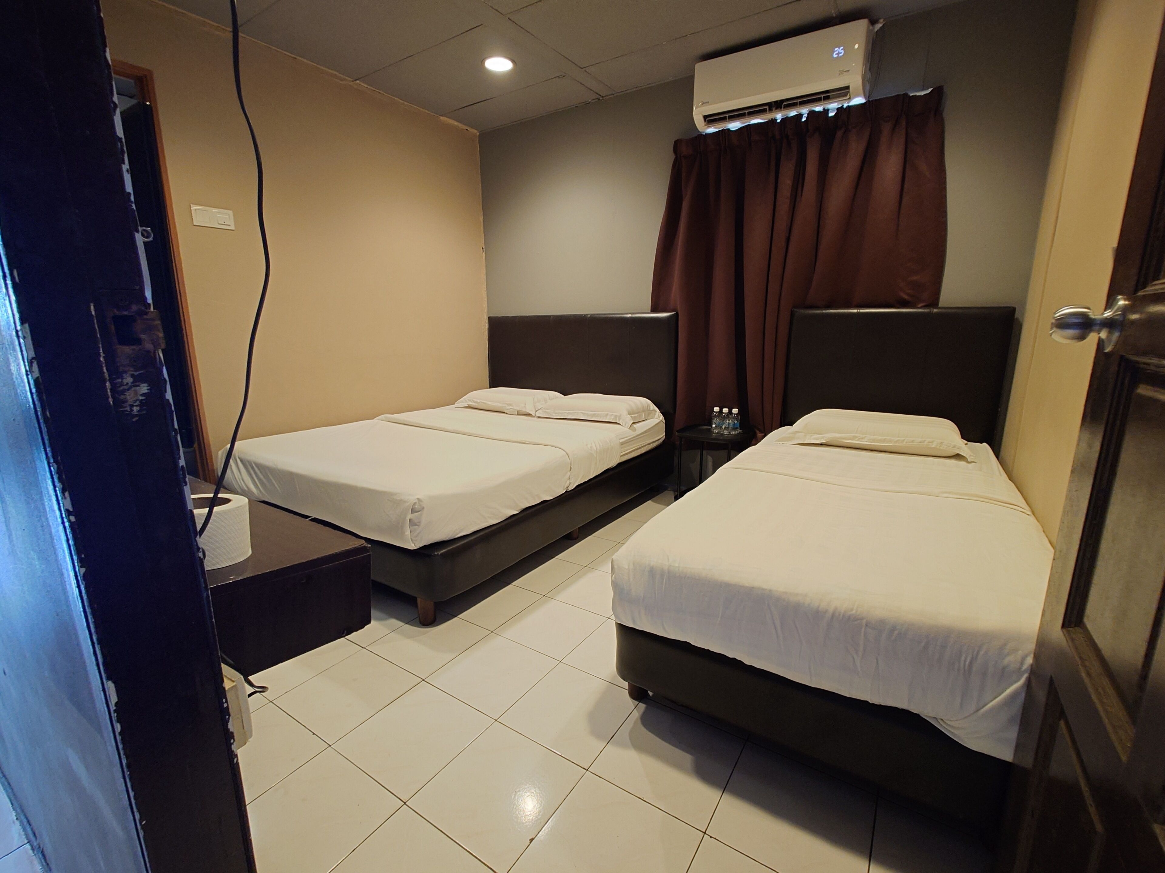 Habitación de hotel con dos camas individuales, una mesita pequeña, una mesita de noche, un equipo de aire acondicionado y una cortina.
