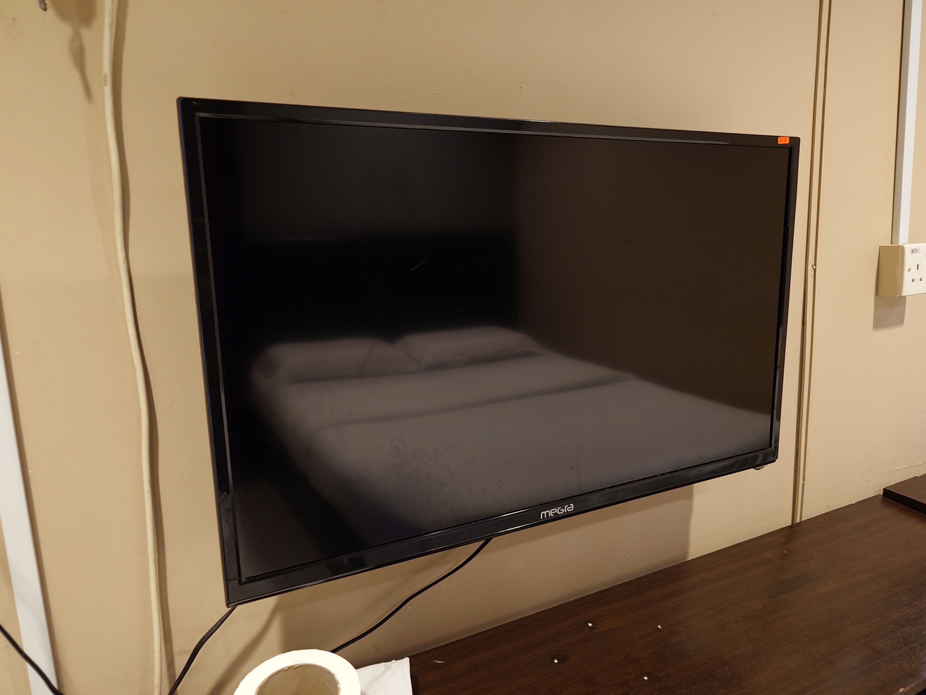 A flat-screen TV mounted on a wall, a wooden desk, and a white power outlet.