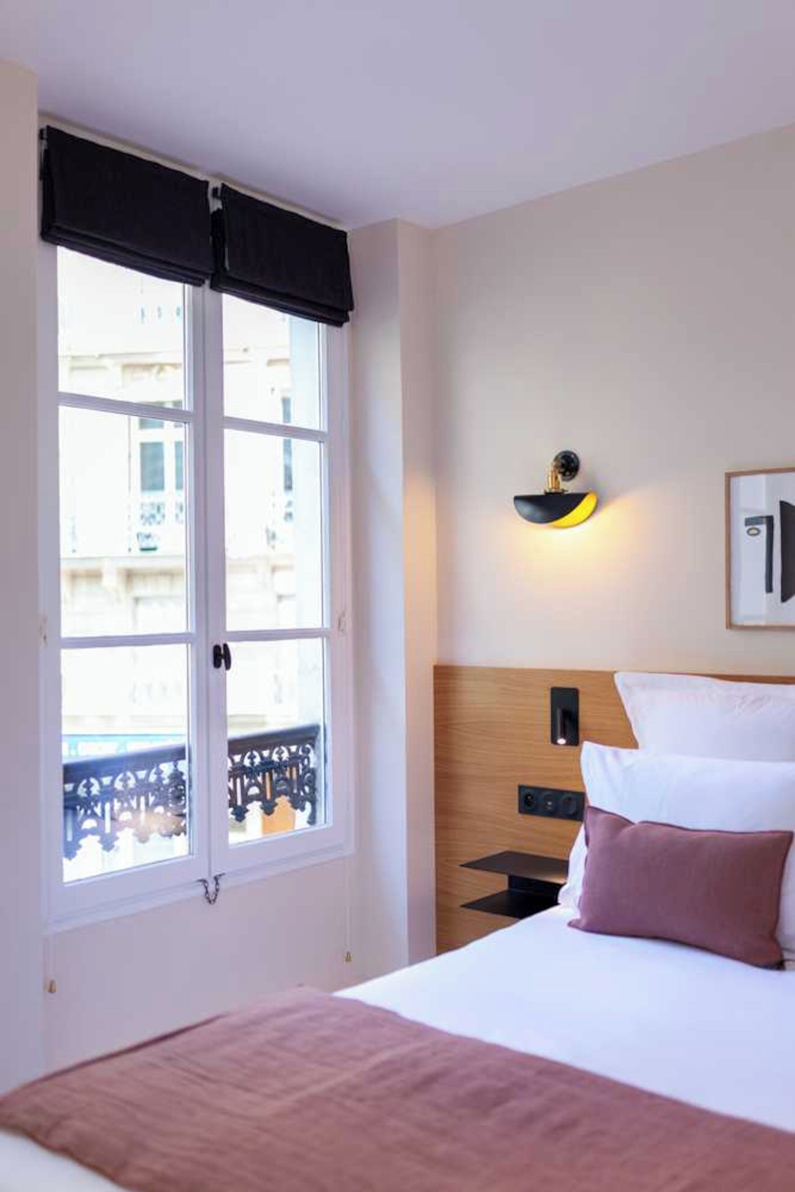 Ein Hotelzimmer mit Bett, Nachttisch, Wandlampe und Fenster mit Blick auf Gebäude.