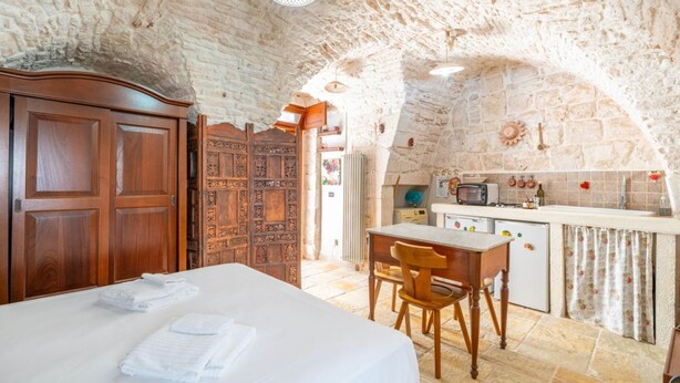 Quarto com cama, guarda-roupa de madeira, escrivaninha com cadeira, pequena cozinha e teto de pedra.