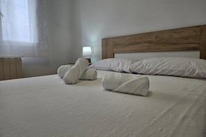 A neatly made bed with rolled towels, a wooden headboard, a bedside lamp, and a window with sheer curtains.