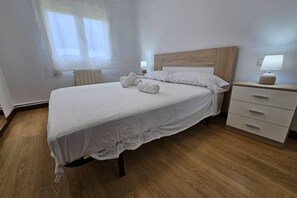 Uma cama bem arrumada com lençóis brancos, cabeceira de madeira, mesa de cabeceira com abajur e janela com cortinas leves.