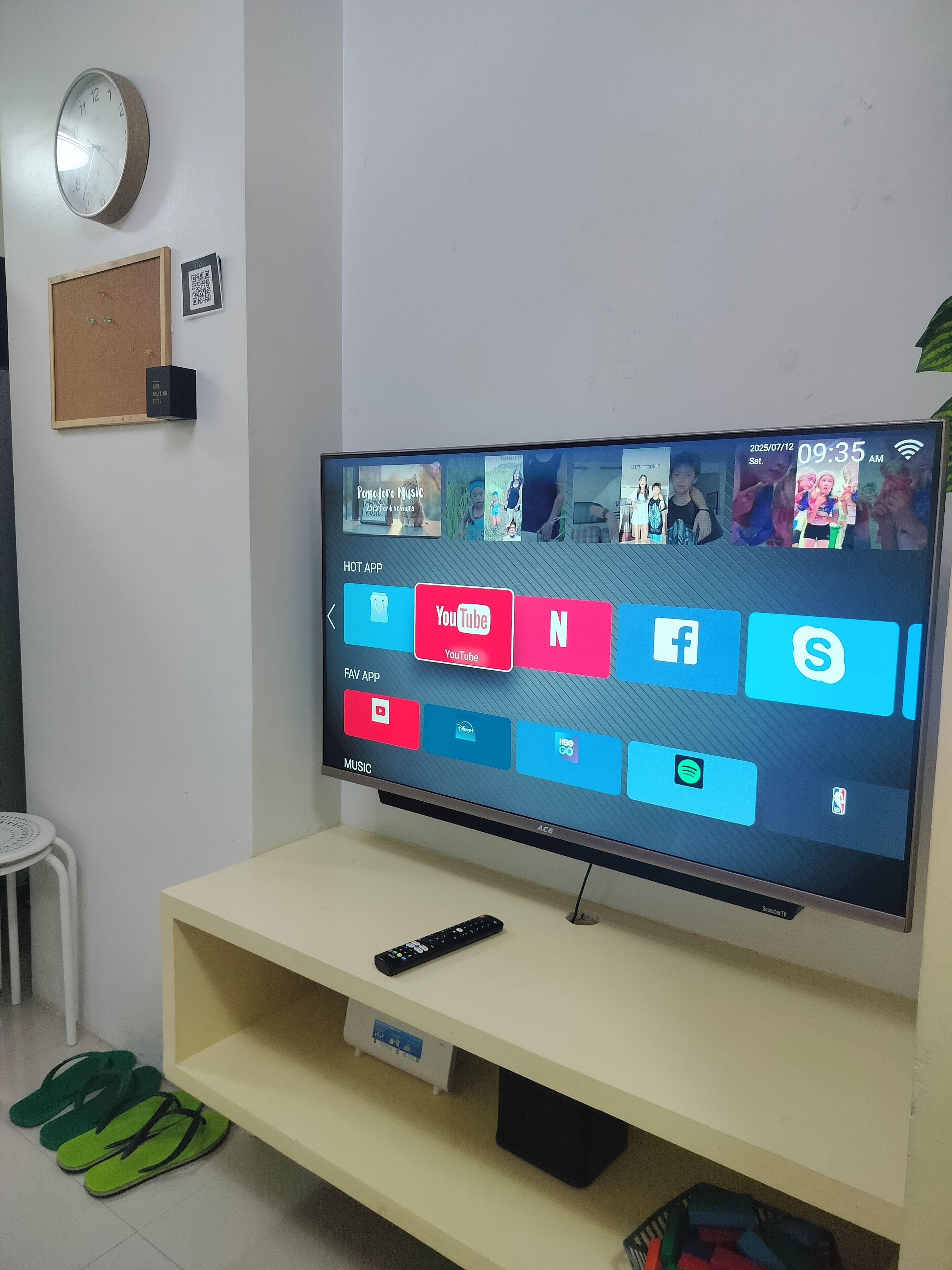 Television, joka on jalustalla, ja sen mukana kaukosäädin, seinäkello ja QR-koodia sisältävä kyltti.