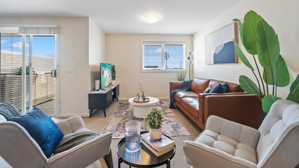 A modern living room with a sofa, armchairs, a coffee table, and a TV. There is a balcony with a view, a painting on the wall, and a large plant.