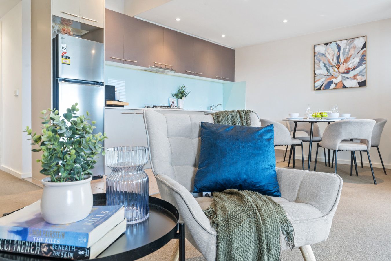 A modern living room with a grey sofa, a blue pillow, a glass vase, and a potted plant. There is a dining area with a table and chairs in the background.