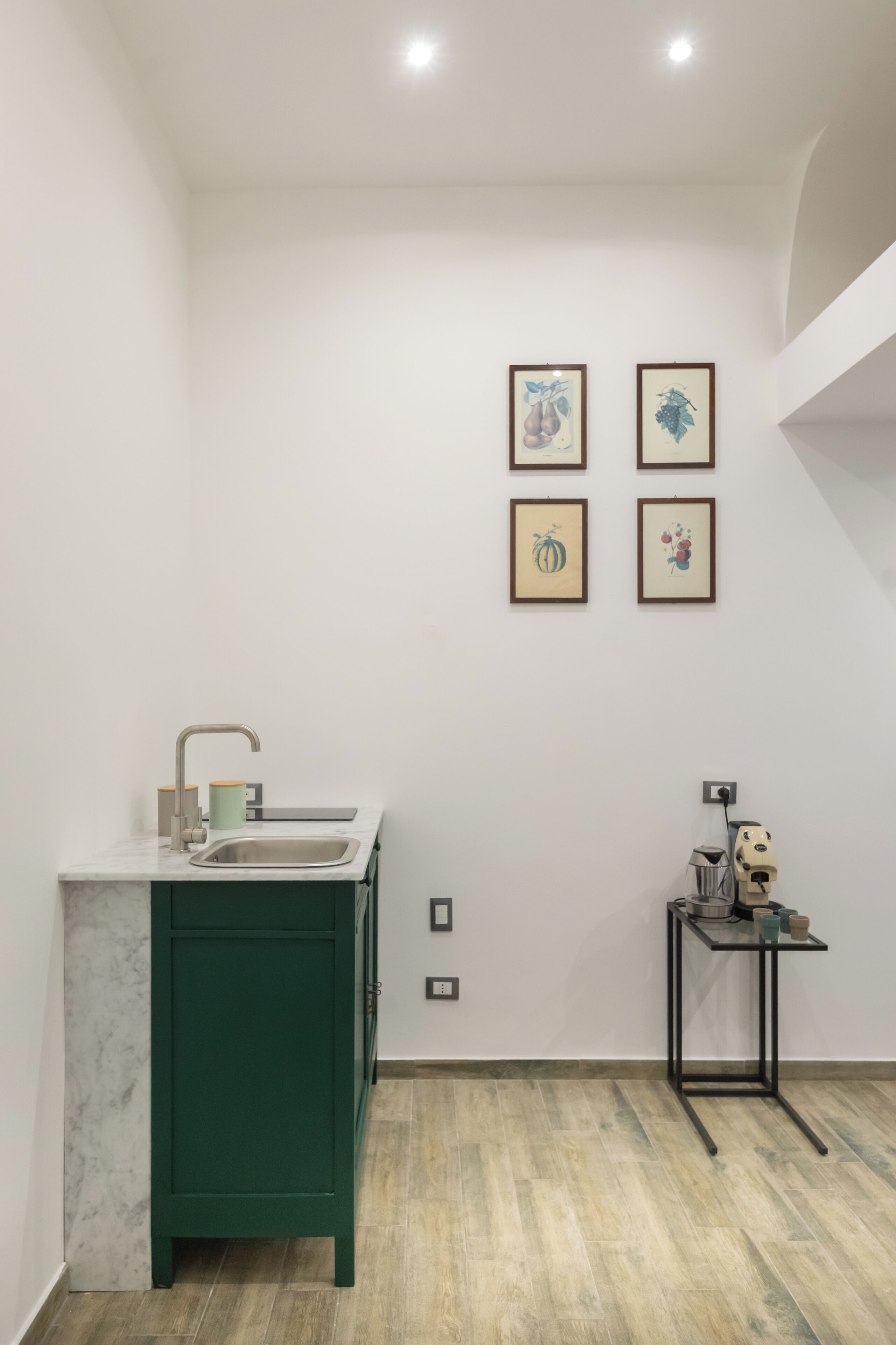 A kitchen with a sink, a green cabinet, and a small table with a lamp. There are four framed pictures on the wall.