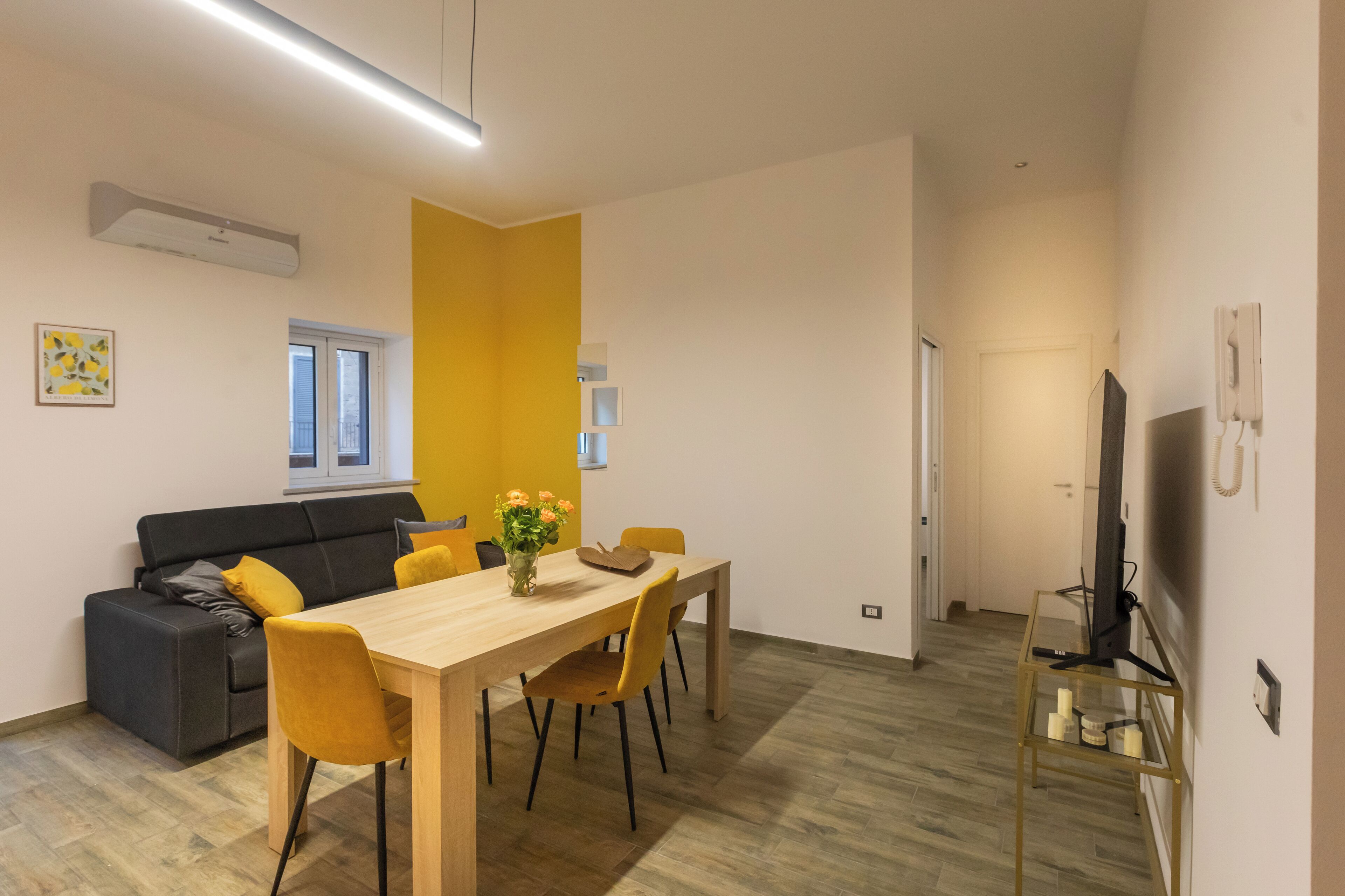 Una sala de estar moderna con una mesa de comedor de madera, sillas amarillas, un sofá gris y un televisor sobre un mueble.