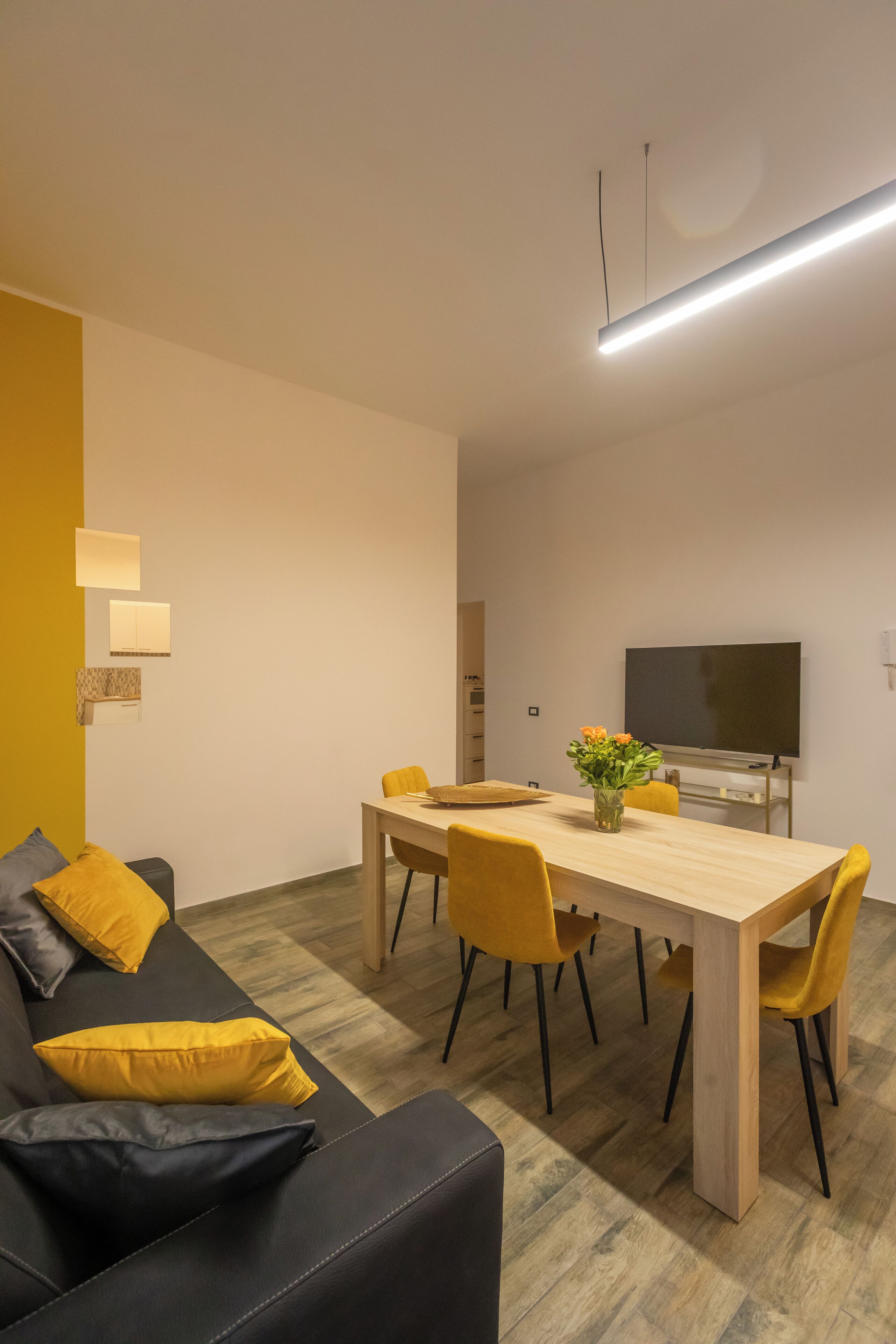 Uma sala de estar moderna com um sofá, uma mesa de jantar e cadeiras. Há uma TV de tela plana em um suporte e um vaso com flores sobre a mesa.