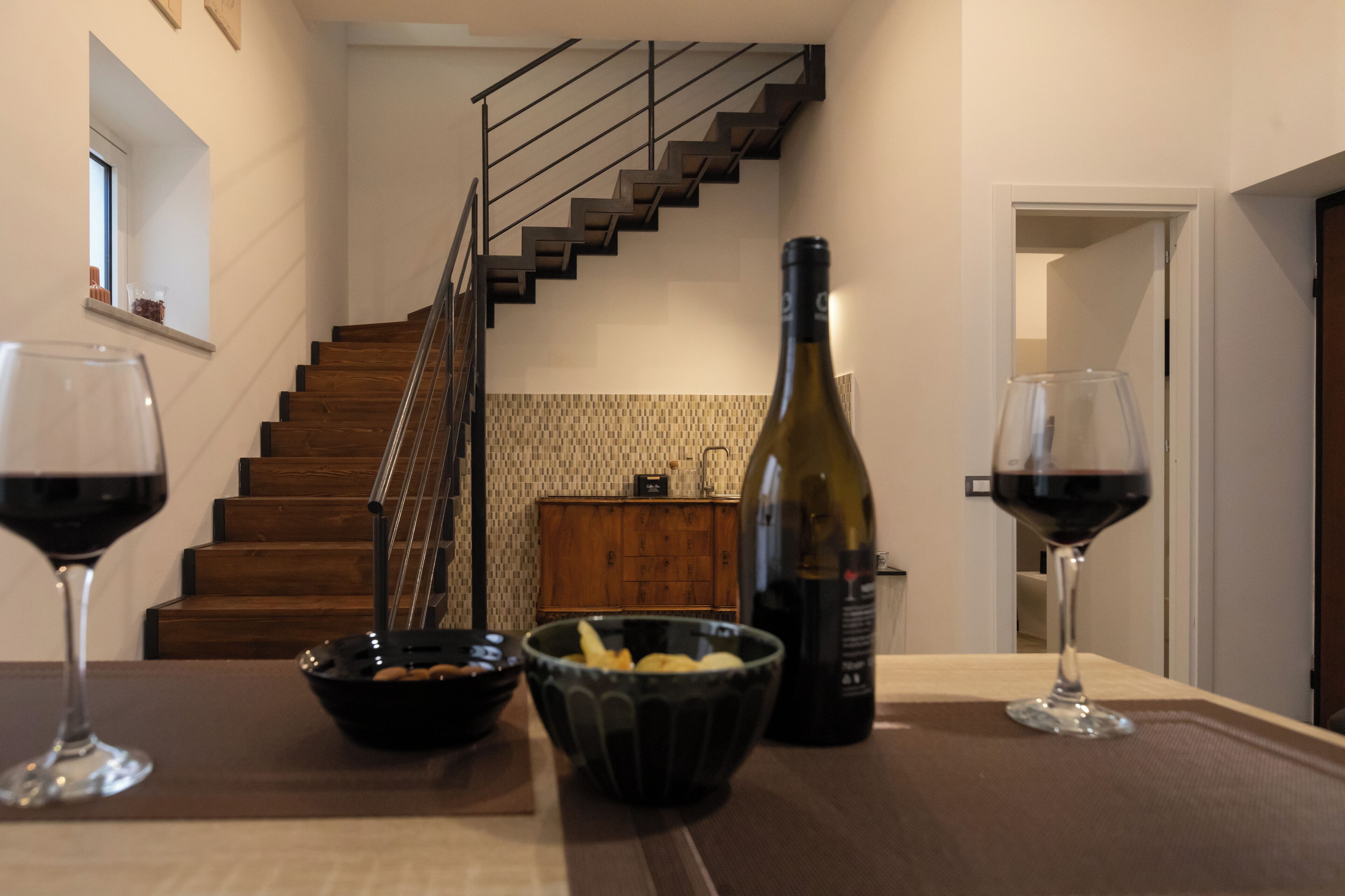 A dining area with a wooden table, two wine glasses, a bowl of snacks, and a bottle of wine. A staircase is visible in the background.