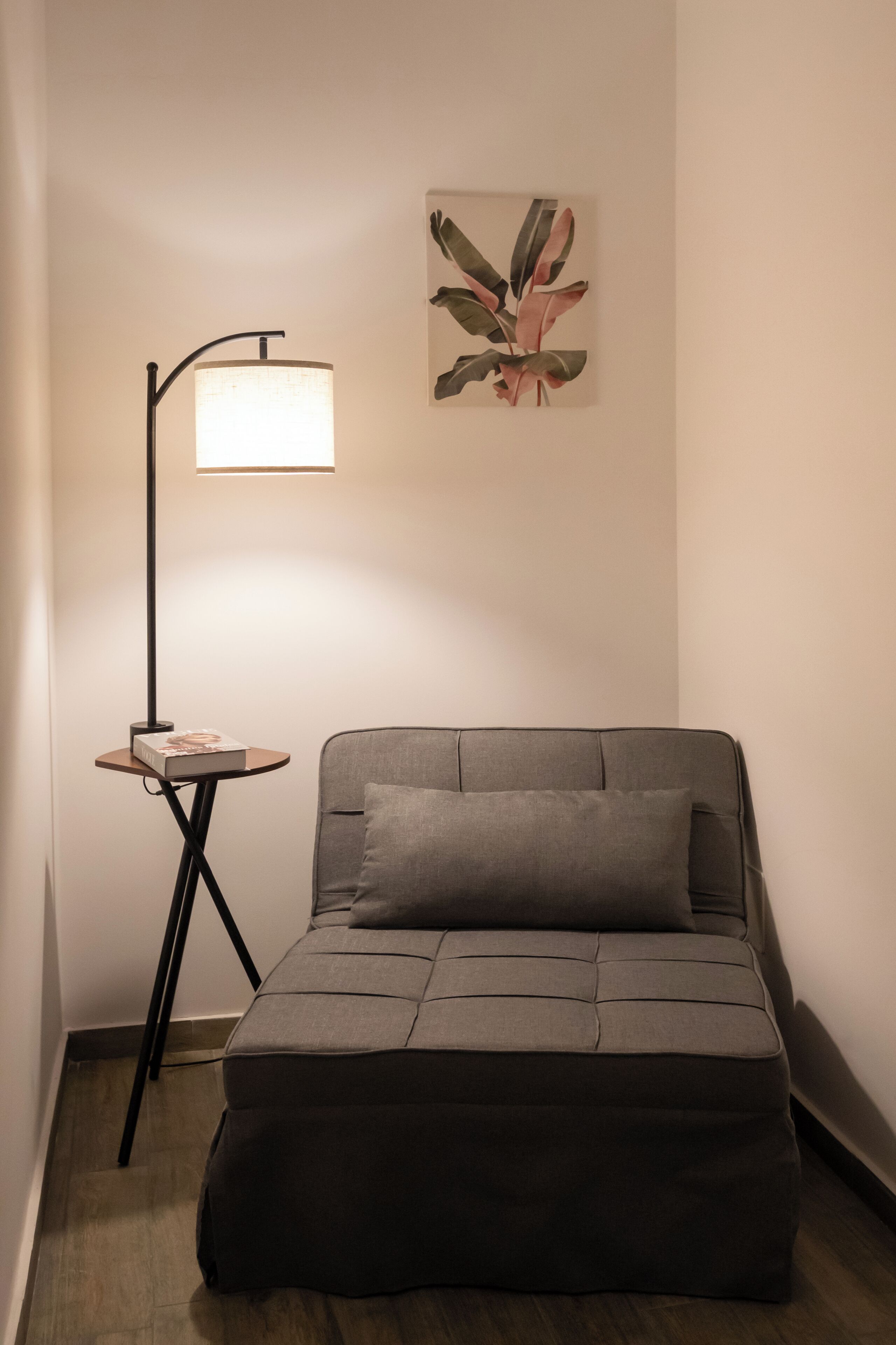 A small, cozy room with a grey sofa, a side table with a lamp, and a framed artwork on the wall.