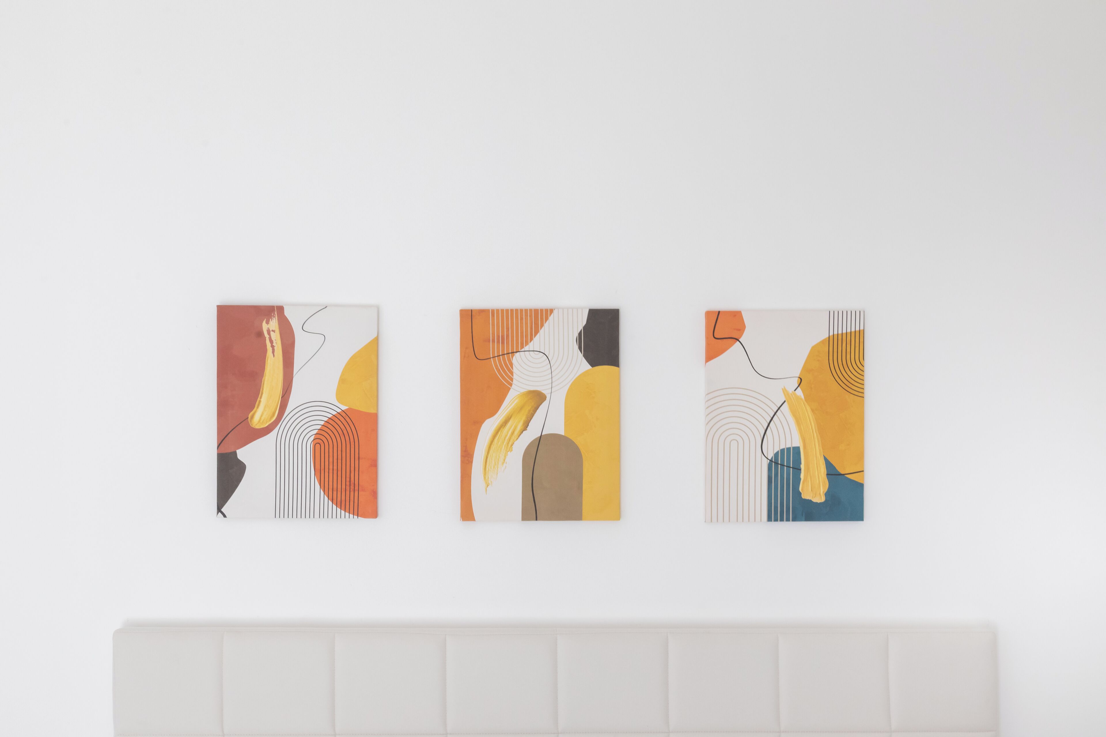 Three abstract paintings on a white wall above a bed with a white headboard.