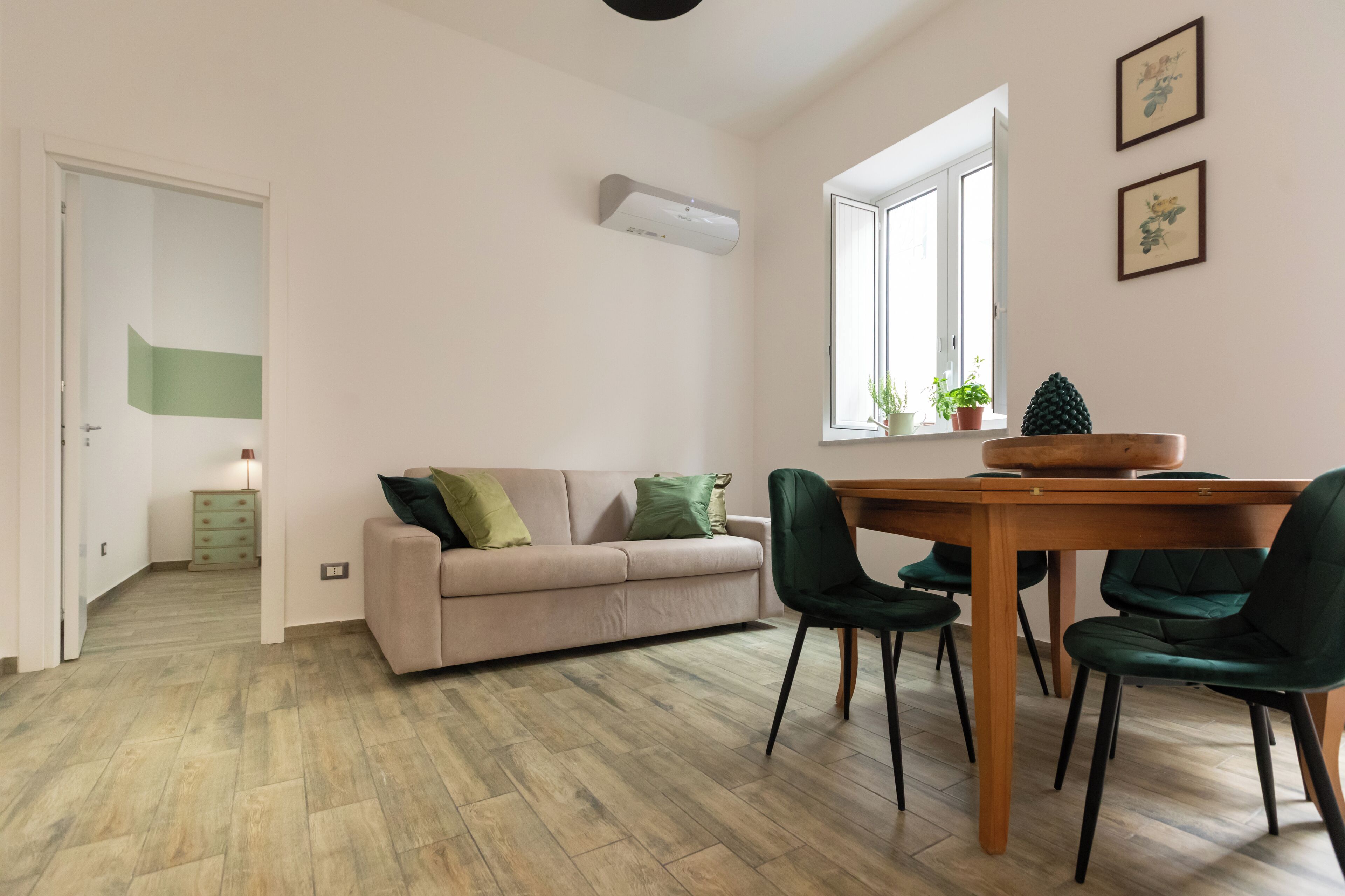 A modern living room with a beige sofa, green cushions, a wooden dining table with chairs, and a window with plants.