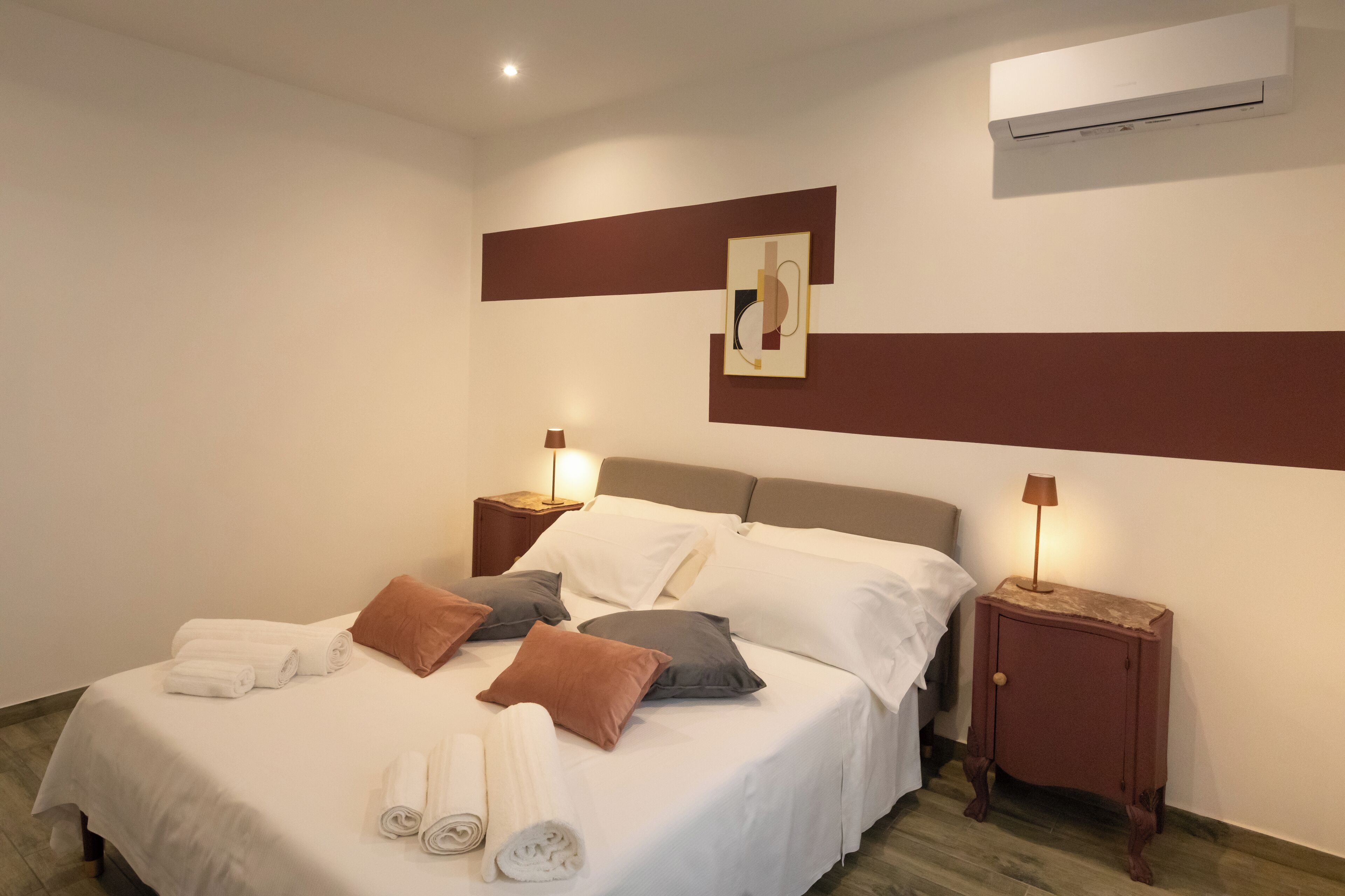 Una cama bien hecha con sábanas blancas y toallas enrolladas, dos mesitas de noche con lámparas, un aire acondicionado de pared y un cuadro enmarcado sobre la cama.
