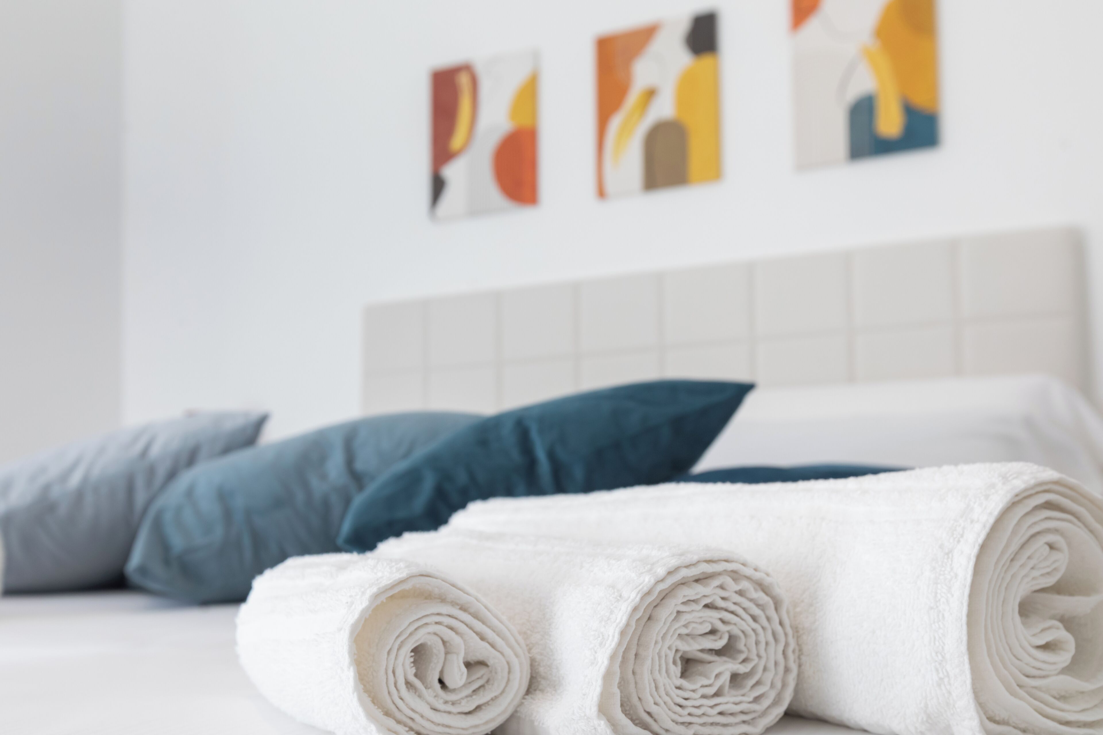A neatly made bed with white and blue pillows, a rolled white towel, and abstract art on the wall.