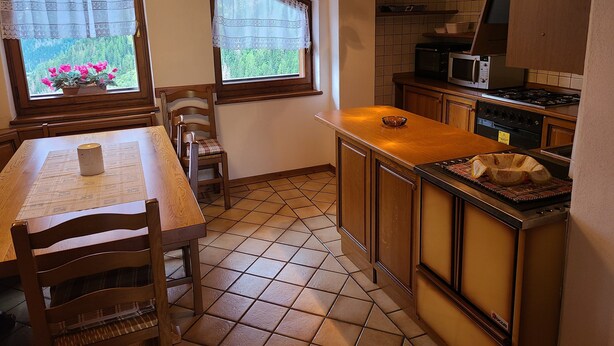 A kitchen with wooden cabinets, a tiled floor, a dining table with chairs, and a window with a view of greenery. - Apartment 'Pozzobon' with Mountain View, Shared Garden and Wi-Fi (Caracoi Agoin)