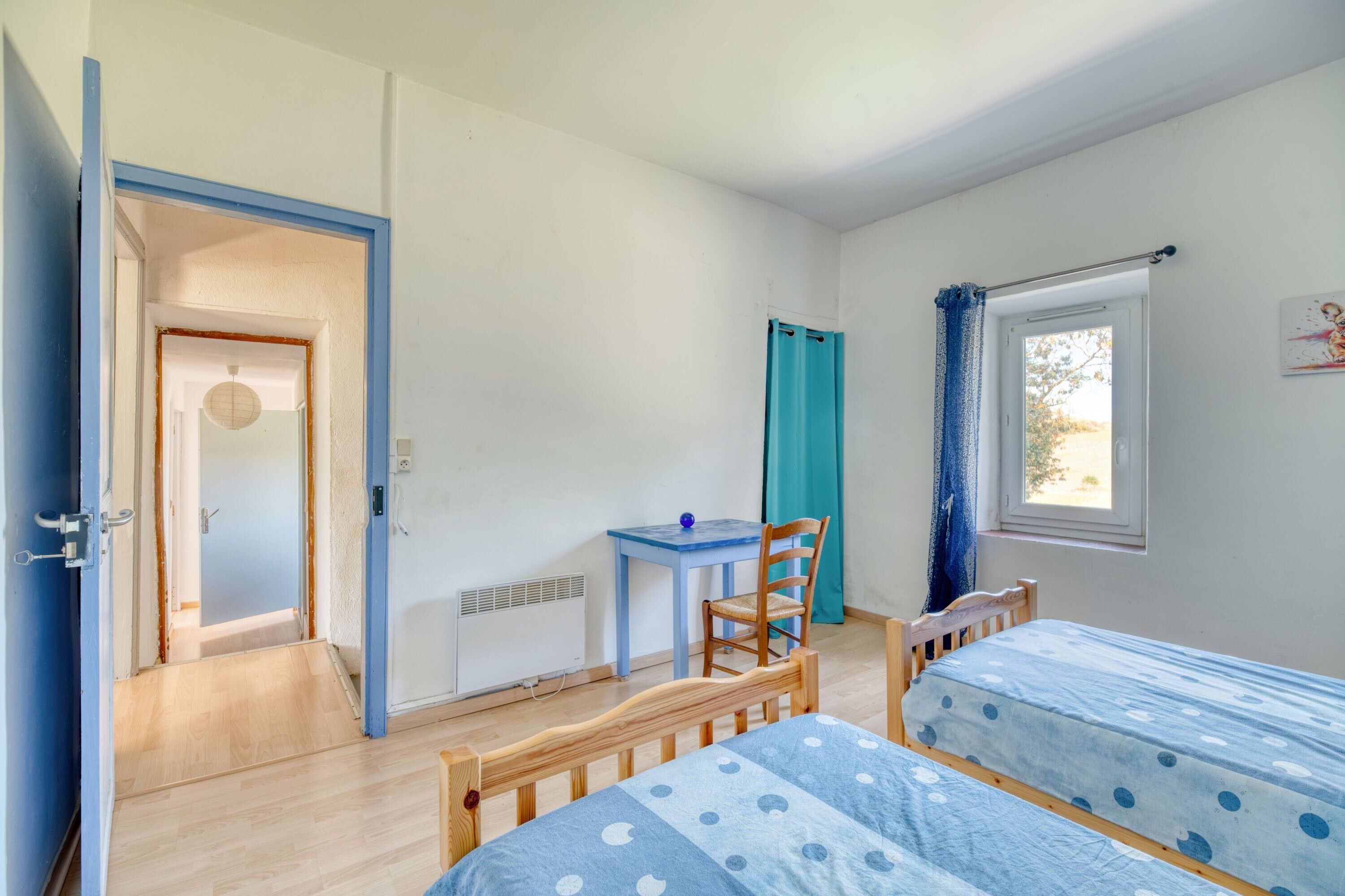 Een kamer met twee bedden, een bureau, een stoel, een raam met gordijnen, en een deur naar een andere kamer.