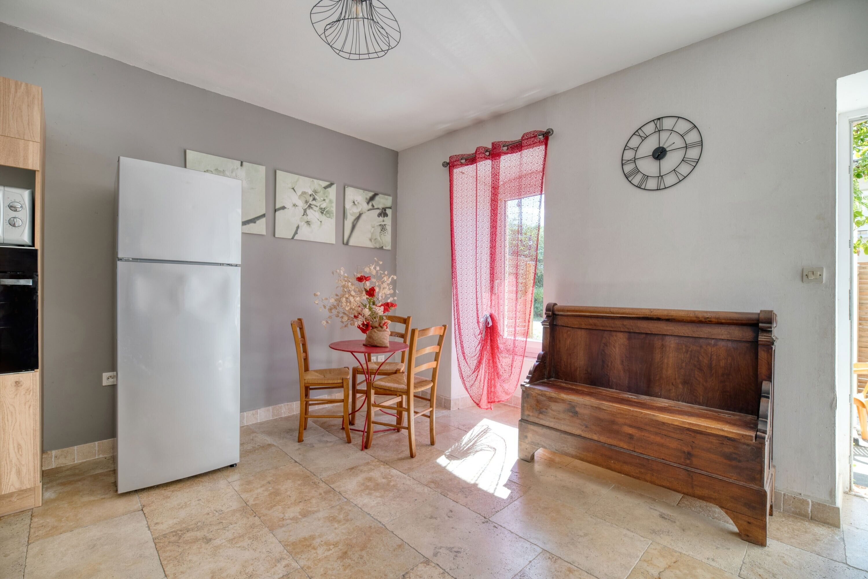 A kitchen with a refrigerator, wooden chairs, a table with flowers, a bench, and wall clocks.