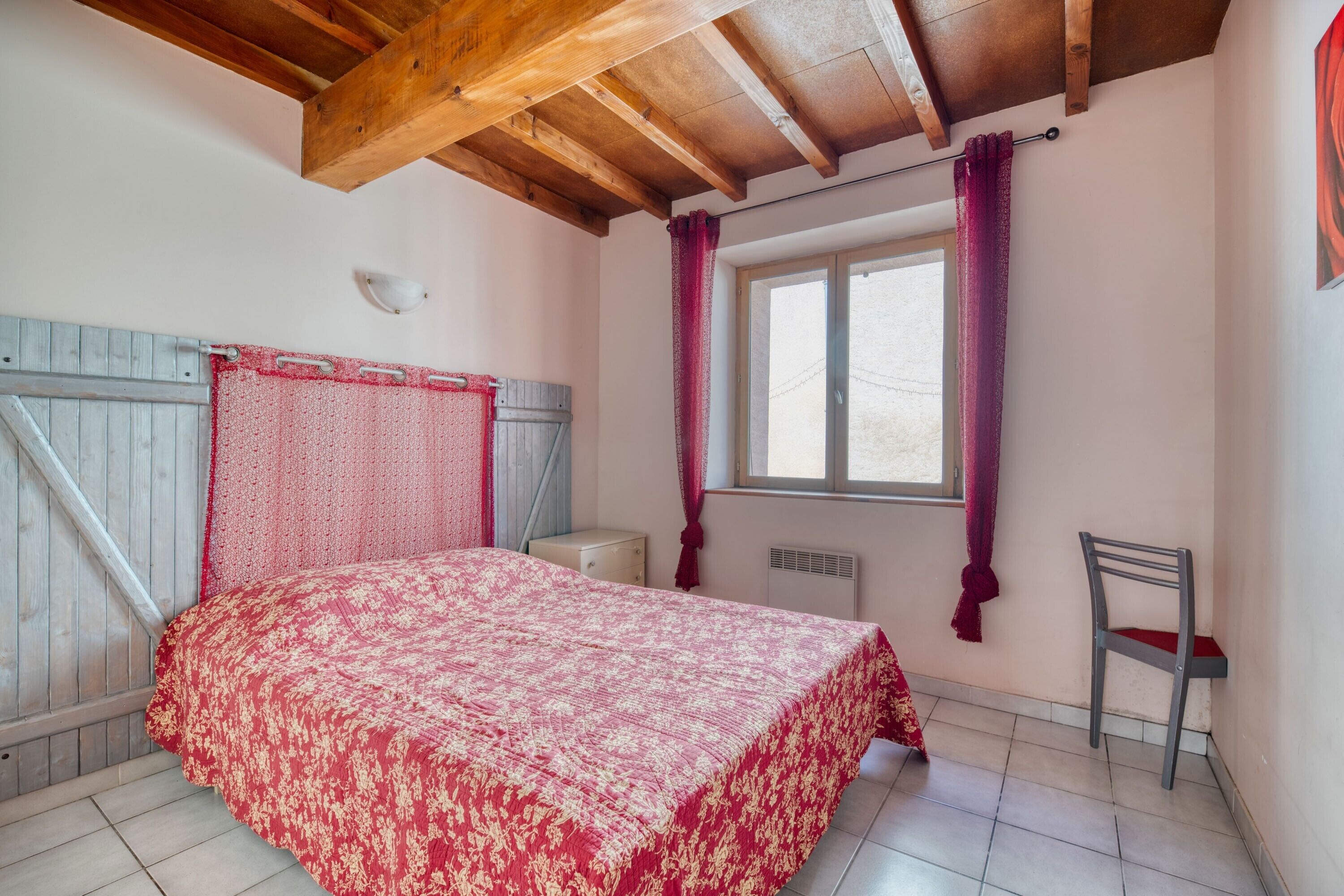 Quarto com teto de madeira, cama com colcha estampada, janela com cortinas, uma cadeira e uma pequena mesa de cabeceira.