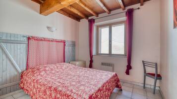 Quarto com teto de madeira, cama com colcha estampada, janela com cortinas, uma cadeira e uma pequena mesa de cabeceira.