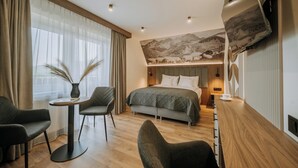 Una habitación de hotel moderna con una cama grande, un área de estar con dos sillones, una mesita con un jarrón y un televisor montado en la pared.