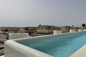 Una piscina con borde de azulejos, rodeada por una valla blanca y con vistas a una zona residencial con casas y palmeras.