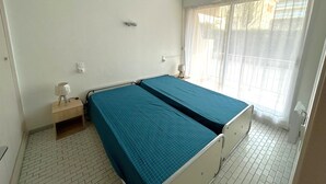 A room with a single bed, a bedside table with a lamp, a window with sheer curtains, and a hanging light fixture. - Apartment 4 guests near beach with parking (Argelès-sur-Mer)