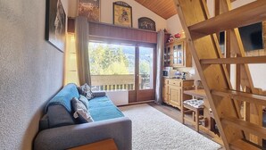 A cozy living room with a sofa, wooden beams, a kitchen area, and a large window with a view. - 2-room duplex with balcony and Wifi facing Mont Blanc (Combloux)