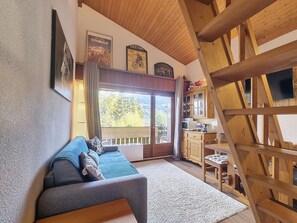 A cozy living room with a sofa, wooden beams, a kitchen area, and a large window with a view. - 2-room duplex with balcony and Wifi facing Mont Blanc (Combloux)