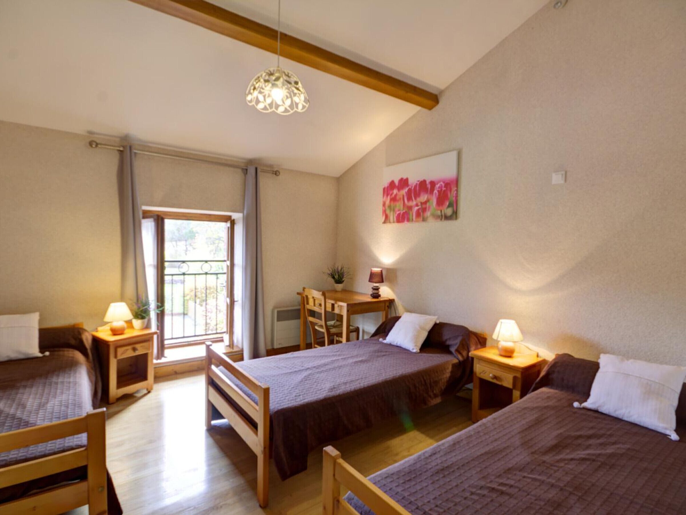 A room with three wooden beds, a desk, and a chair. There is a window with curtains, a hanging light fixture, and a painting on the wall.
