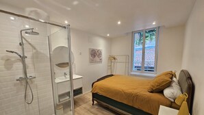 Une chambre moderne dotée d’une cabine de douche en verre, d’un lit avec une couvre-lit de couleur moutarde, d’une table de chevet avec une lampe et d’une fenêtre donnant sur un mur de briques.