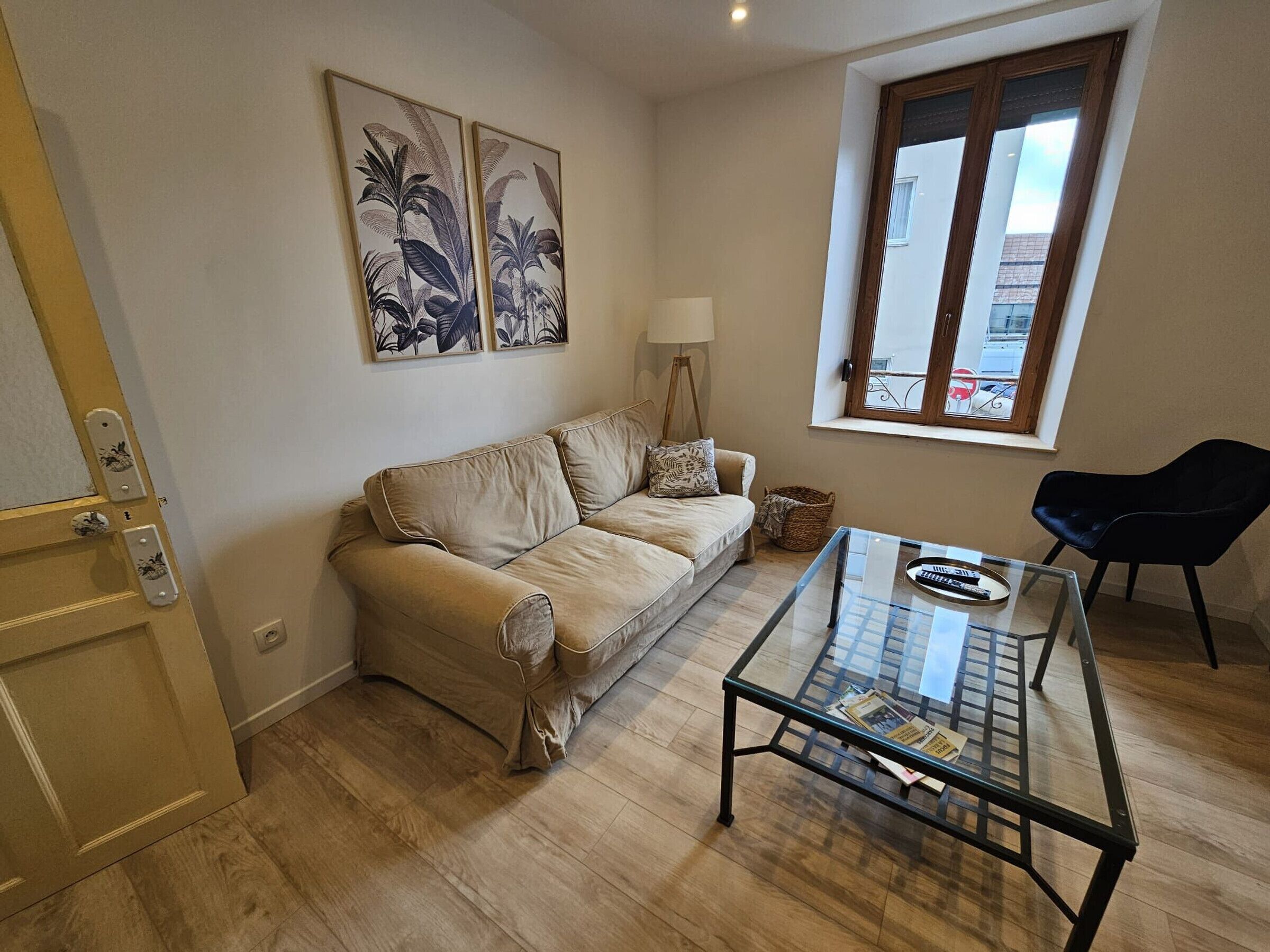 A living room with a beige sofa, a glass coffee table, a black chair, a window with a view, and two wall-mounted paintings.