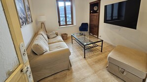 Un salon avec un canapé beige, une table basse en verre, un fauteuil bleu, un meuble en bois et un téléviseur à écran plat fixé au mur.
