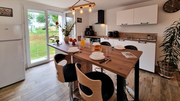 Une cuisine moderne avec une table en bois, quatre chaises, un luminaire suspendu et une vue sur l’extérieur.