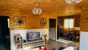 A cozy living room with a wooden ceiling, a television, a sofa, and a dining area with a table and chairs.