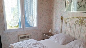 Une chambre à coucher avec un lit, une table de chevet avec une lampe, une fenêtre avec des rideaux en dentelle et un papier peint à motifs floraux.