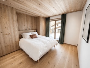 A bedroom with a wooden ceiling, a bed with white bedding, and a view through a glass door.