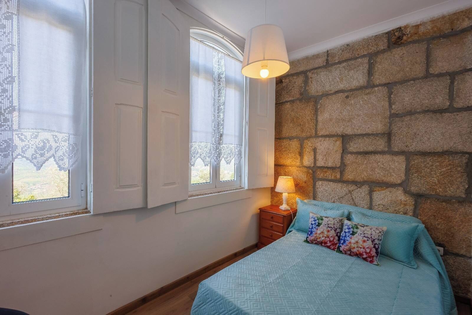 A bedroom with a bed, a nightstand with a lamp, and a window with lace curtains.